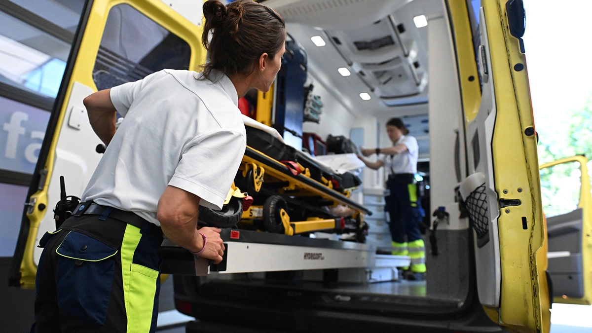 Une ambulancière charge un brancard dans une ambulance, tandis qu'un collègue prépare l'équipement à l'intérieur de l'ambulance.