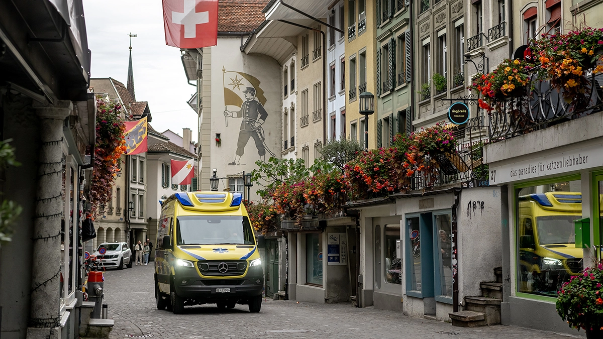 Une ambulance traverse la vieille ville historique avec le drapeau suisse et des façades ornées de fleurs, services de secours en intervention urbaine.