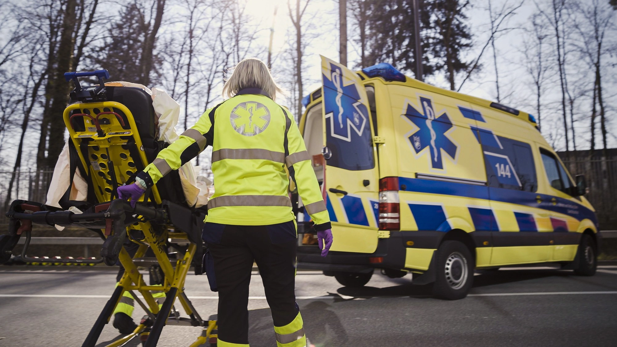 Une ambulancière charge un brancard dans une ambulance à la lumière du jour, intervention des services de secours sur une route ouverte.