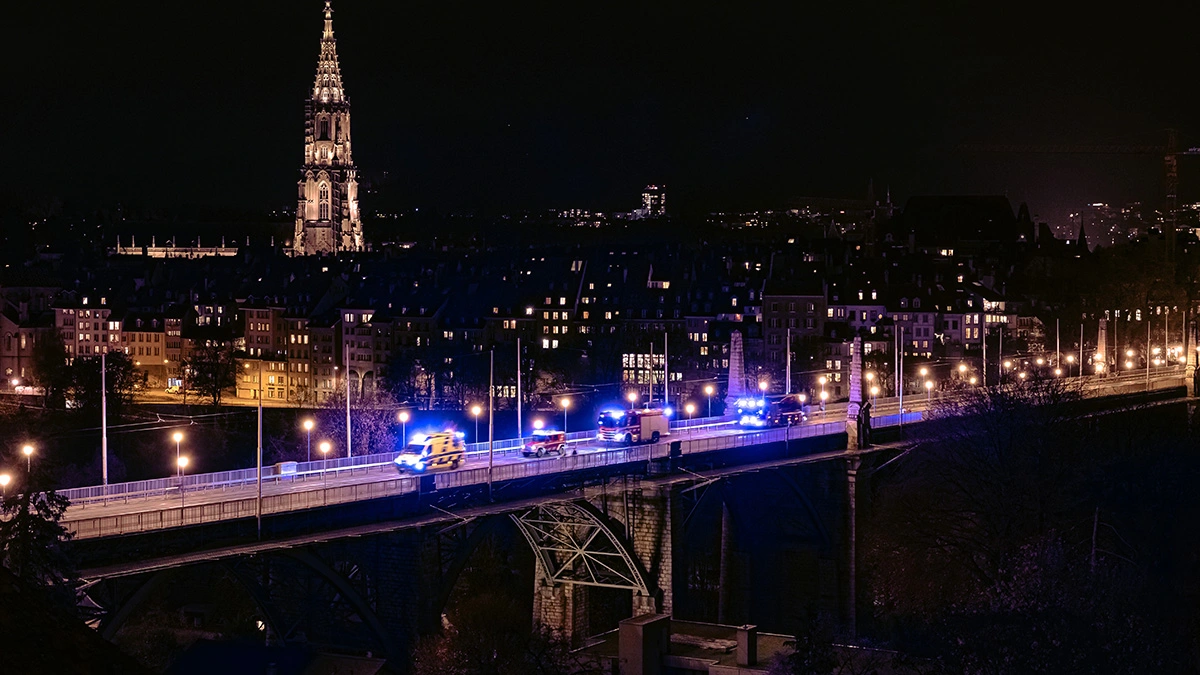 Une ambulance avec gyrophare roule de nuit sur le pont illuminé de la Kornhausbrücke à Berne, intervention de sauvetage devant la toile de fond de la cathédrale de Berne.